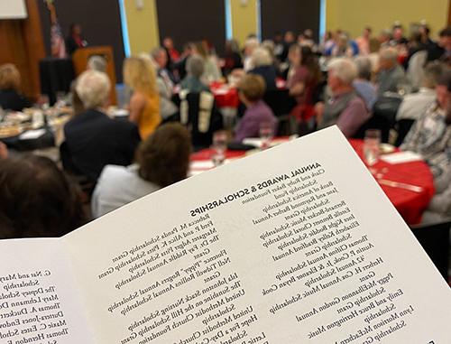 A scholarship luncheon program in the foreground with a group of guests in the background